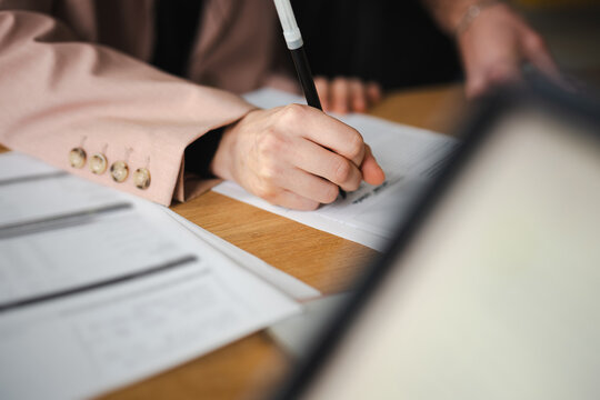 A person in a blazer writes on a document with a pen, surrounded by papers and a tablet on a wooden desk.