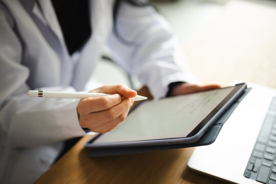 A person in a white coat uses a stylus to interact with a tablet, with a laptop nearby.