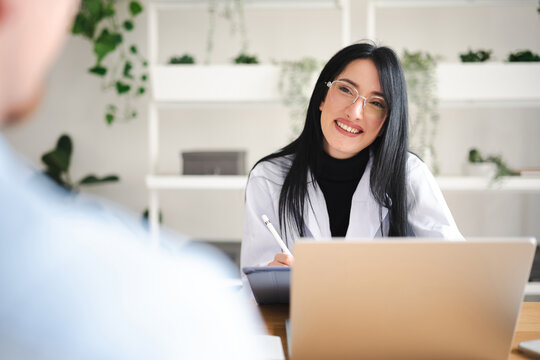 A smiling woman in a white coat and glasses is working on a laptop and tablet, interacting with a patient in a bright, modern office setting.