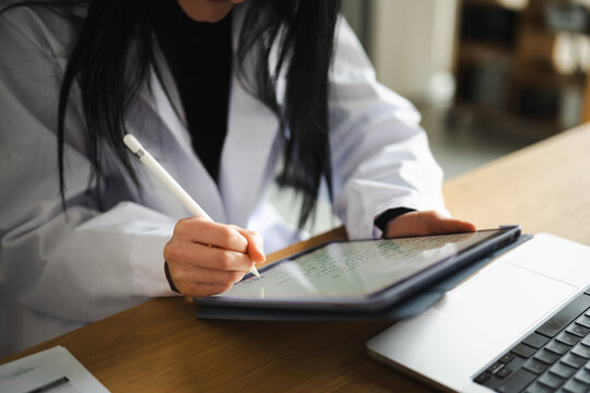 A person in a white lab coat uses a stylus to write on a tablet, with a laptop nearby on a wooden desk.