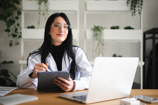 A woman in a white lab coat and glasses works on a tablet with a stylus, with a laptop open on her desk.