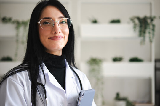 A smiling female doctor wearing glasses and a white coat holds a tablet, ready to assist patients with care and expertise.