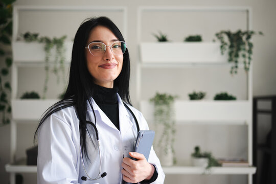 A smiling female doctor in a white coat and stethoscope holds a tablet, ready to assist patients in a modern, plant-filled office.