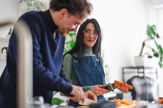 A man and a woman are in a kitchen preparing food together. The woman is peeling a carrot while the man is slicing something on a cutting board.