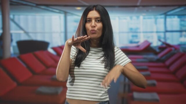 Woman posing with hands under chin and on hip while puckering lips in airport lounge with red reclining seats and floor to ceiling windows; playful affection.