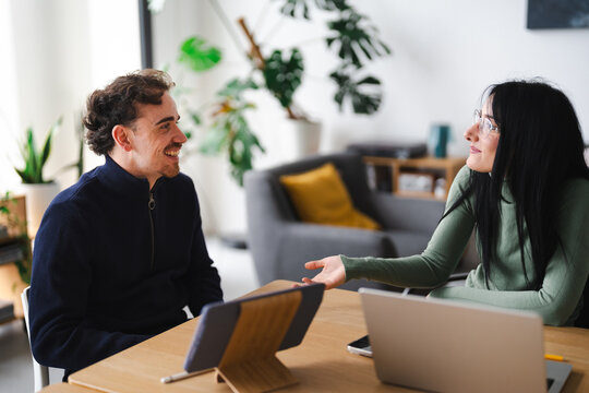 A man and a woman are engaged in a conversation at a table with a laptop and tablet. They are smiling and gesturing, suggesting a collaborative discussion.