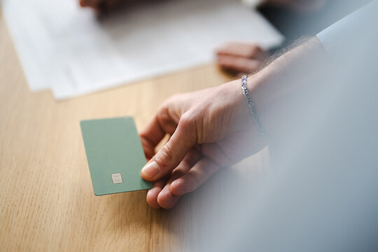 A person's hand holds a green payment card over a wooden surface, with blurred documents in the background.
