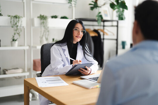 A doctor in a white coat consults with a patient, taking notes on a tablet in a modern office setting.