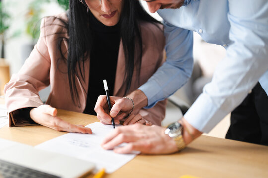 A man and woman collaborate at a desk, with the woman signing a document while the man offers guidance.