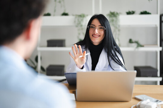 A smiling doctor in a white coat gestures with her hand while speaking to a patient in a modern office setting.