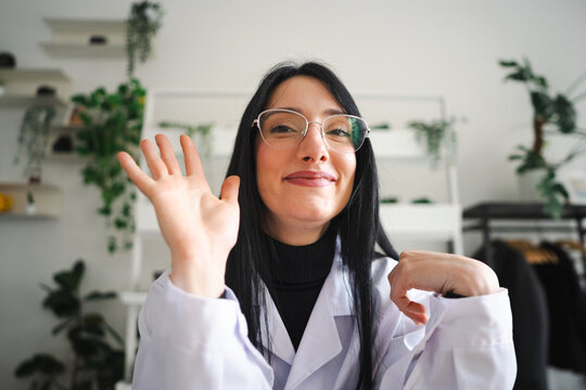 A smiling woman wearing glasses and a lab coat waves hello. She is in a bright room with plants in the background.