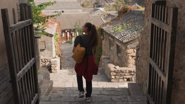 Asian woman with yellow backpack descending stone steps through open wooden gate, polkadot top, red jacket tied waist, back view, warm lateafternoon light, trailing ivy, quiet heritage lane, steady