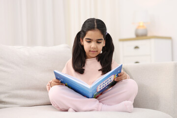 Naklejka premium Cute little girl reading book on couch at home