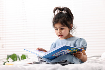 Naklejka premium Cute little girl reading book on bed at home. Space for text