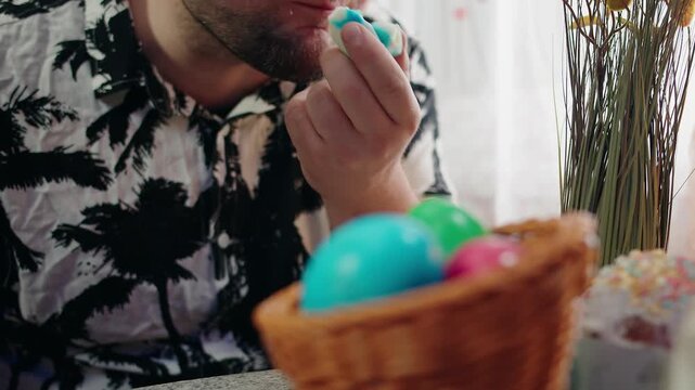 Bearded adult caucasian man holding and eating colorful painted easter egg. Wicker basket with dyed eggs on kitchen table. Festive dinner and celebration of the spring holiday. No face