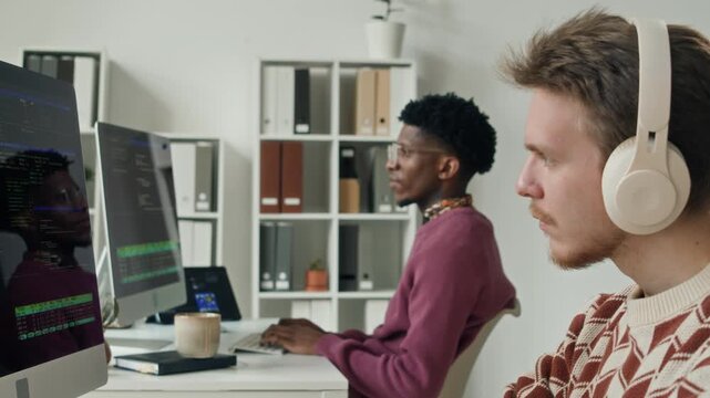 Rack focus of young male programmer with headset on coding at workstation beside young Black teammate in modern open workspace