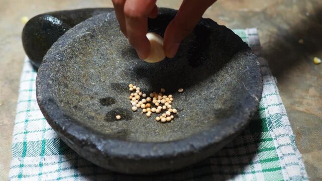 Hand adding coriander, garlic, candlenut, and turmeric to a traditional stone mortar, preparing spices for cooking