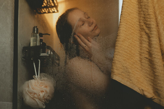 Young woman practicing self-care ritual in cozy bathroom, washing hair behind glass shower door with water drops. Morning routine, authentic lifestyle