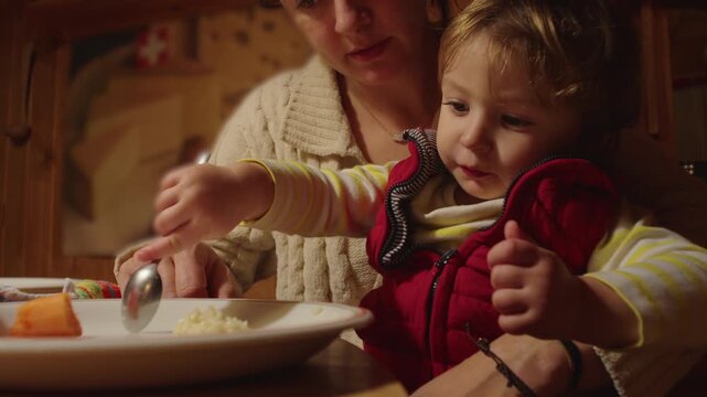 Toddler eating with spoon while mother gently supports hand at wooden dining table inside cozy alpine chalet during family dinner