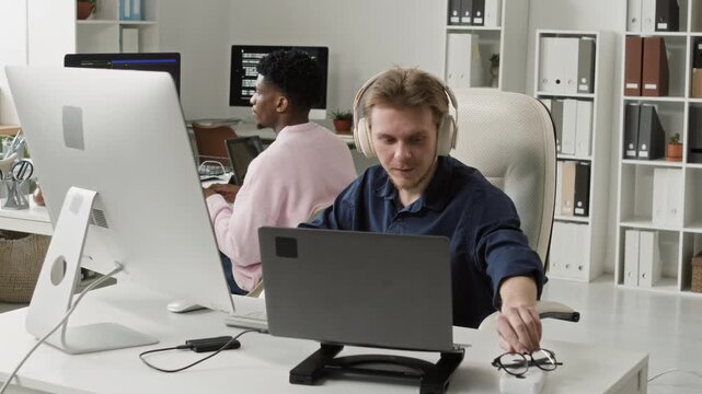 Tired young Caucasian male programmer removing eyeglasses then stretching out arms after long computer coding while sitting at desk with laptop in open space office with colleagues