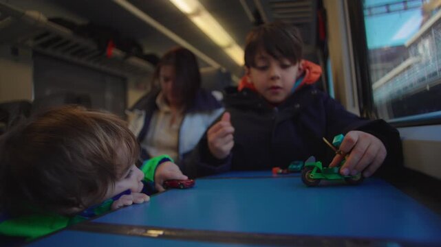 Two brothers playing with toy cars and figures on train table during family rail journey, mother seated behind while urban landscape passes outside window