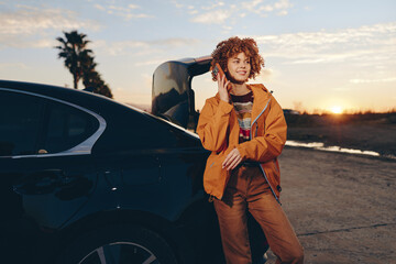 Young woman with curly hair wearing rainbow sweater and orange jacket stands by black car trunk outdoors at sunset smiling and talking on smartphone enjoying lifestyle moment. © SHOTPRIME STUDIO