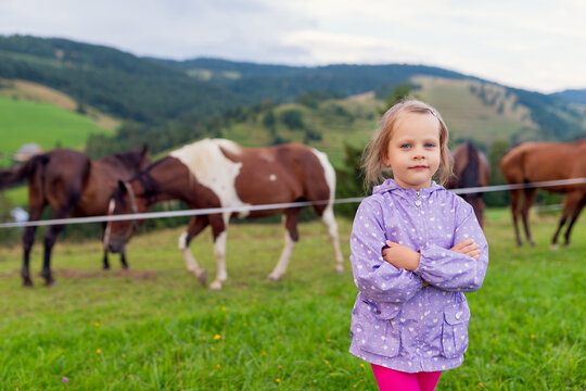 Little girl in purple jacket stands with arms crossed in green mountain pasture, horses grazing in background, rural countryside childhood portrait with copy space.