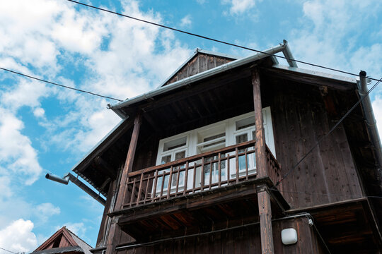 Rustic wooden house facade with balcony and white windows, low angle view under blue sky with clouds, traditional countryside cabin architecture.