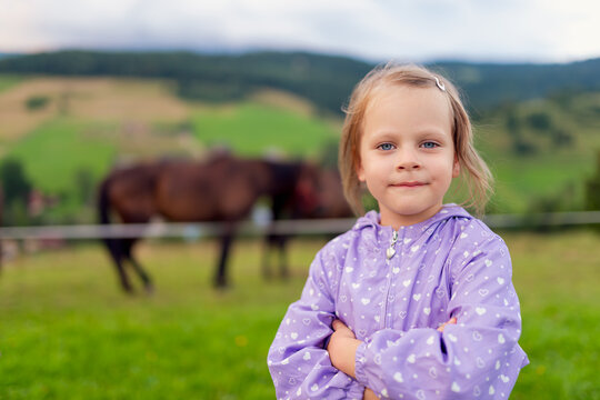 Little girl in purple rain jacket with arms crossed in mountain pasture, horses blurred in background, rural countryside child portrait with copy space.