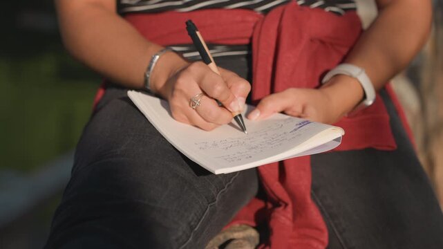 Closeup hands writing notebook outdoors, student drafts lecture notes, poet composes lines, traveler records route, counselor scribbles observations warm sunlight, denim lap, red sweater tied, silver