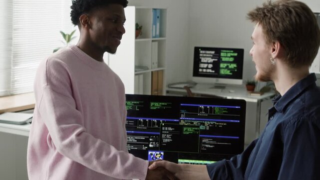 Tilt up shot of young Black male IT programmer shaking hands with coworker confirming code bug fix while standing at computer desk in modern shared office space
