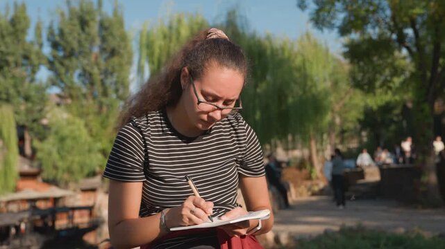 Young woman with curly hair writes notes outside. Student with backpack studies in sunny campus environment. Casual female student with glasses and curly hair focuses on note taking outdoors
