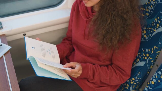 Woman reviewing workbook in train seat, hands turning pages and tracing notes, cozy red hoodie and glasses, window light and passing fields, calm focused practice and quiet study ritual on a long