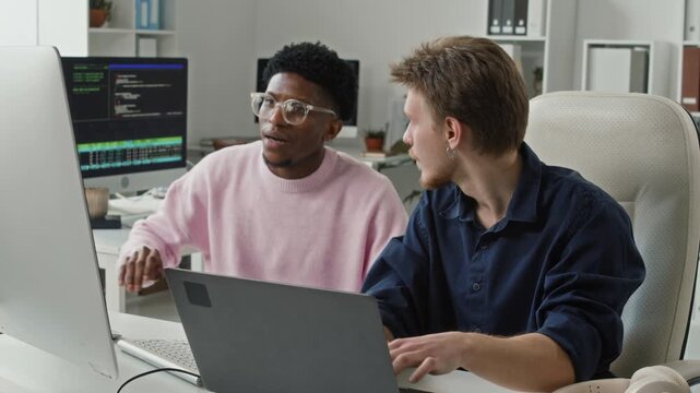Young adult Black male IT specialist coming to desk of senior developer asking question about code optimisation while collaborating on task in modern open plan office