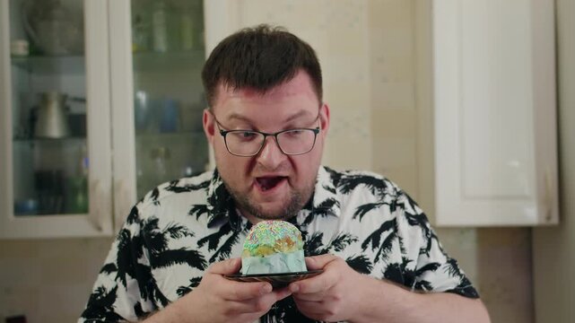 Caucasian bearded man with glasses, shirt and surprised expression sniffing easter cake with glaze and sprinkles. Tasting reaction and comic emotion on face in cozy kitchen. Pampering and fun