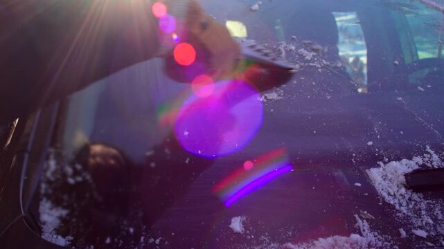 Gloved hand clearing frost from car windshield with child reflection visible in glass, bright winter sunlight and snow around parked vehicle