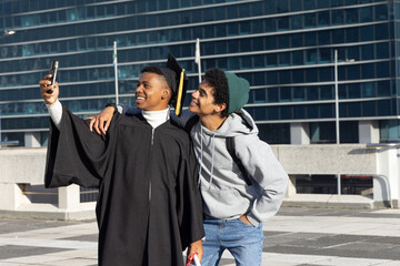 Diverse male friends taking selfie on terrace by glass building with gown cap diploma smartphone © wavebreak3