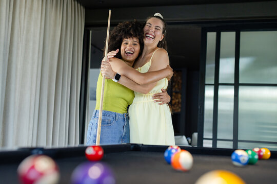 Diverse female friends laughing and hugging in game room with pool table cue and billiard balls