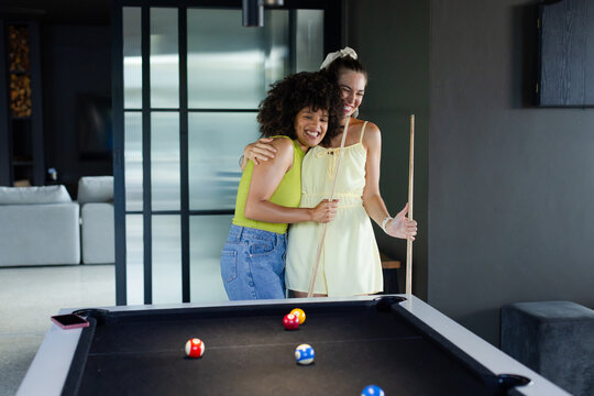 Diverse female friends hugging in game room at black-felt pool table holding pool cue and balls