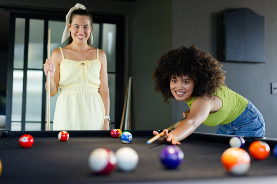 Diverse female friends leaning over pool table in home game room lining up shot with cues