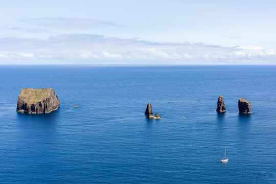 Rock formations in Atlantic Ocean off San Miguel Island coast, Azores, Portugal. Jagged rocks stand tall in blue water. Small sailboat passing by in background.