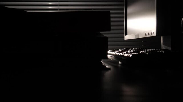 Obscure computer monitor and keyboard on a desk, illuminated by light filtering through horizontal blinds, creating a moody, mysterious atmosphere after hours