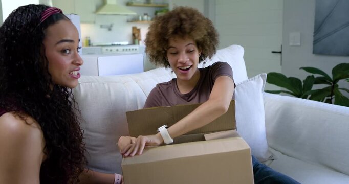 Two African American women opening sealed box on couch, revealing bubble wrap, unwrapping appliance