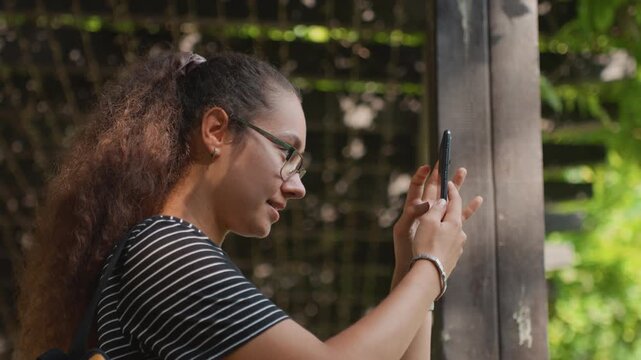 Young woman in striped shirt focused on smartphone near wooden post, capturing plant texture and light quiet contemplative mood, student researcher and amateur photographer roles, gentle bokeh