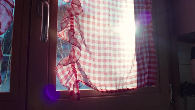 Sunlight shining through red gingham kitchen curtain with visible window handle, rustic home detail evoking warmth and peaceful countryside lifestyle