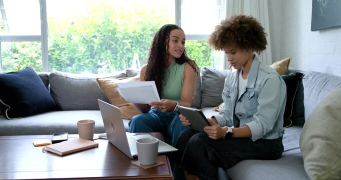 Two African American women starting with papers and tablet on sofa, flipping, using laptop, working