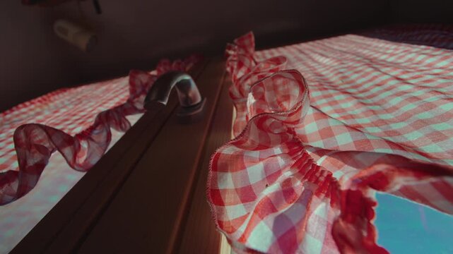Red gingham curtain glowing in sunlight over chalet window, low angle view of checkered fabric and metal handle against bright blue sky