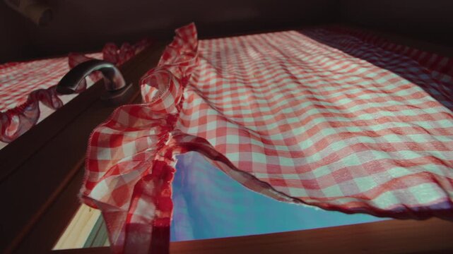 Low angle view of red and white checkered curtains filtering sunlight inside alpine chalet, wooden window frame and handle visible