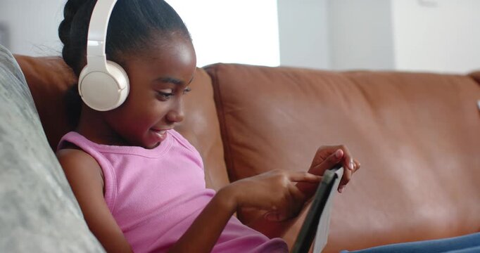 African American girl reclining on sofa wearing pink top, white headphones, tapping tablet learning