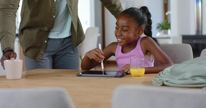 African American dad approaching with mug, leaning and supervising girl drawing on tablet at table